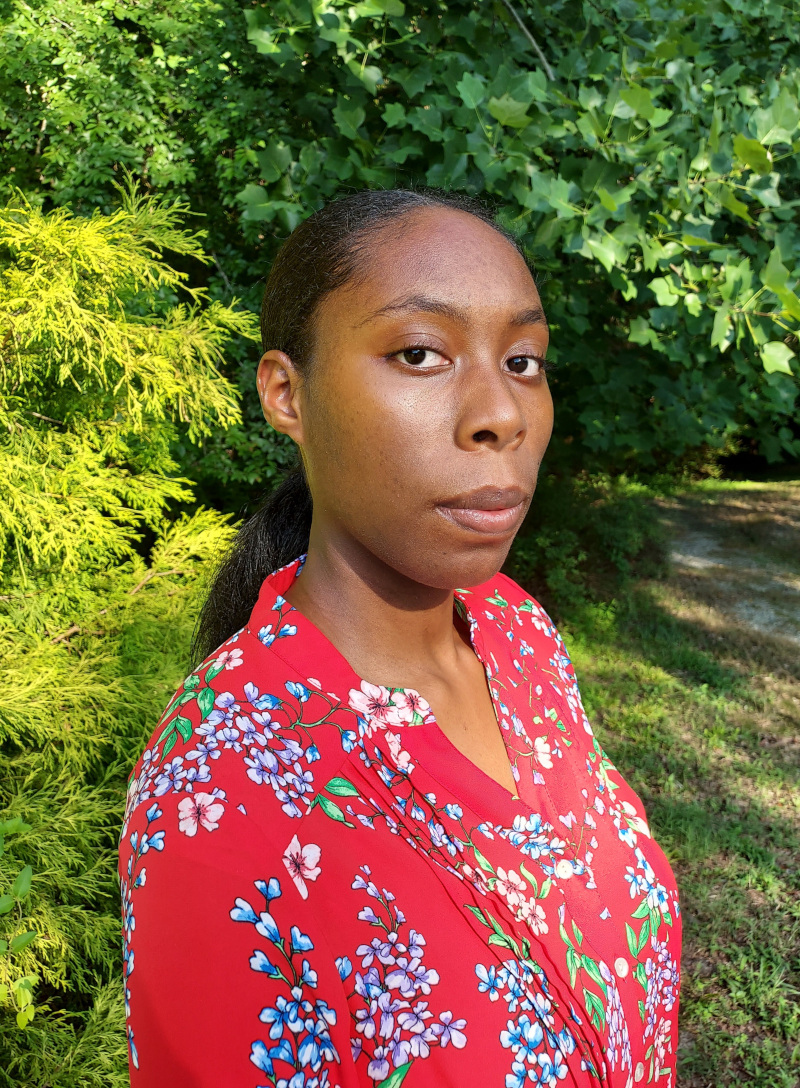 Jasmine, a Black American woman with her black hair tied back in a ponytail, is standing against a backdrop of evergreen and deciduous foliage. She is visible from the chest up and is wearing a vibrant red blouse with a floral pattern. Though she is not smiling, she is looking warmly at the camera.