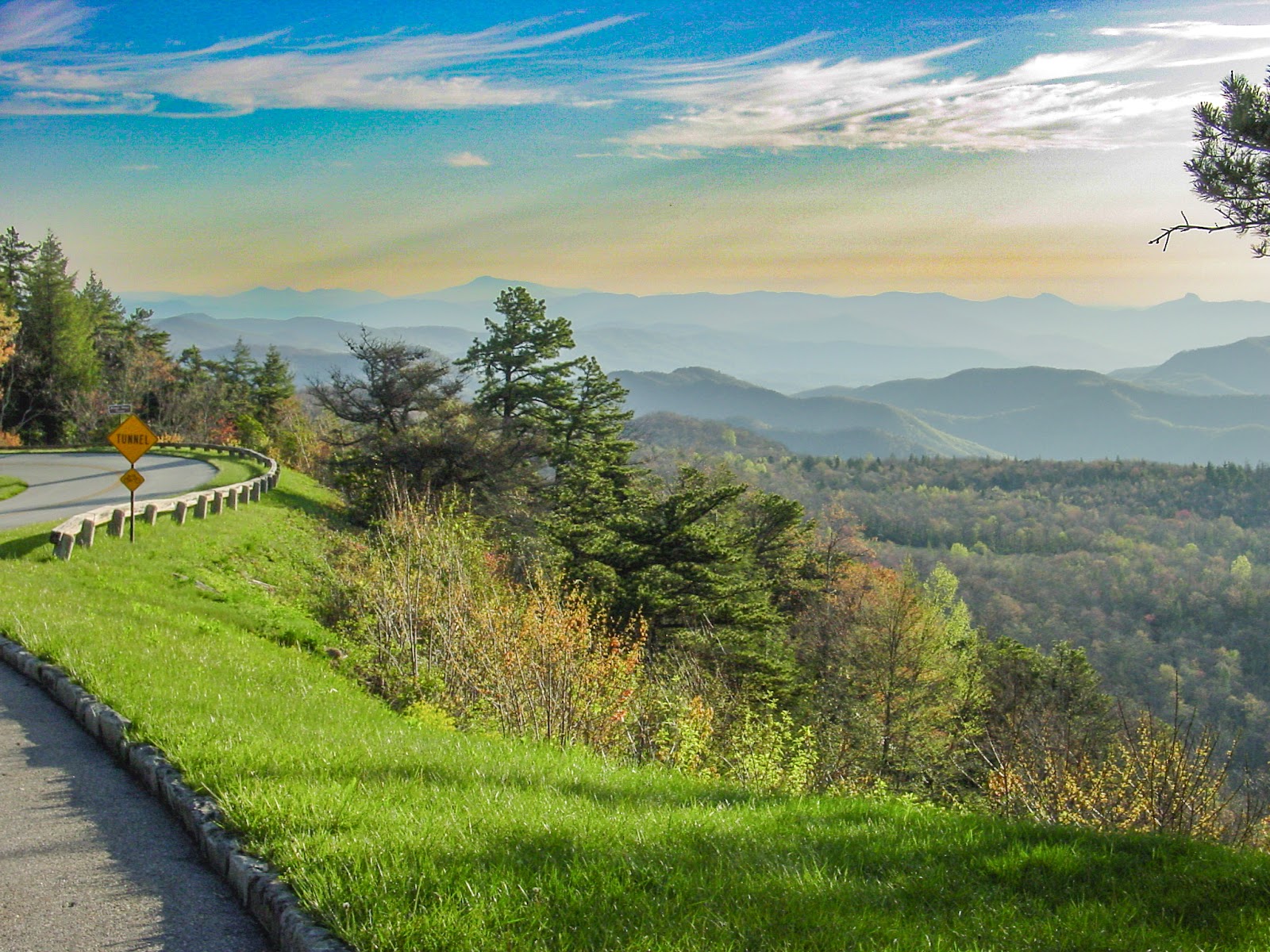 A curvy road on the left opens up to a valley and high mountains and a light orange sky that fades into blue clouds