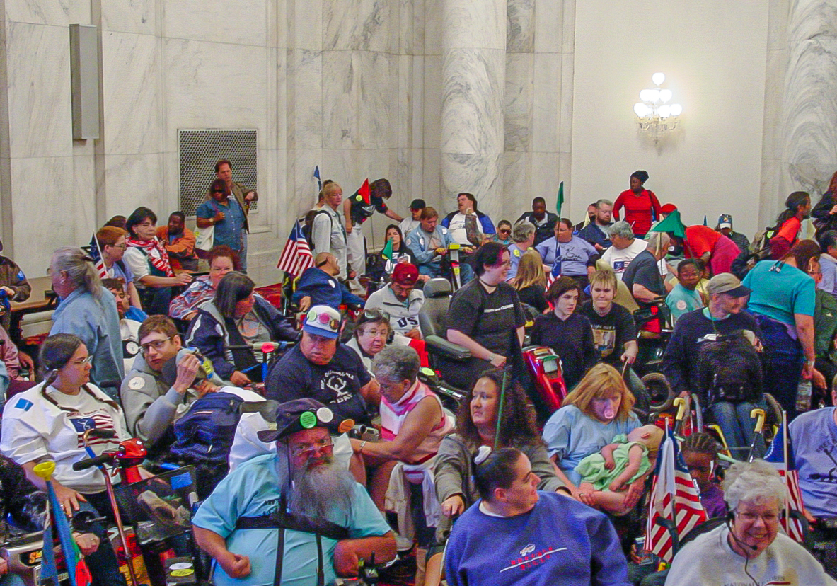Colorful swarm of people in a white marble room with high ceilings. 