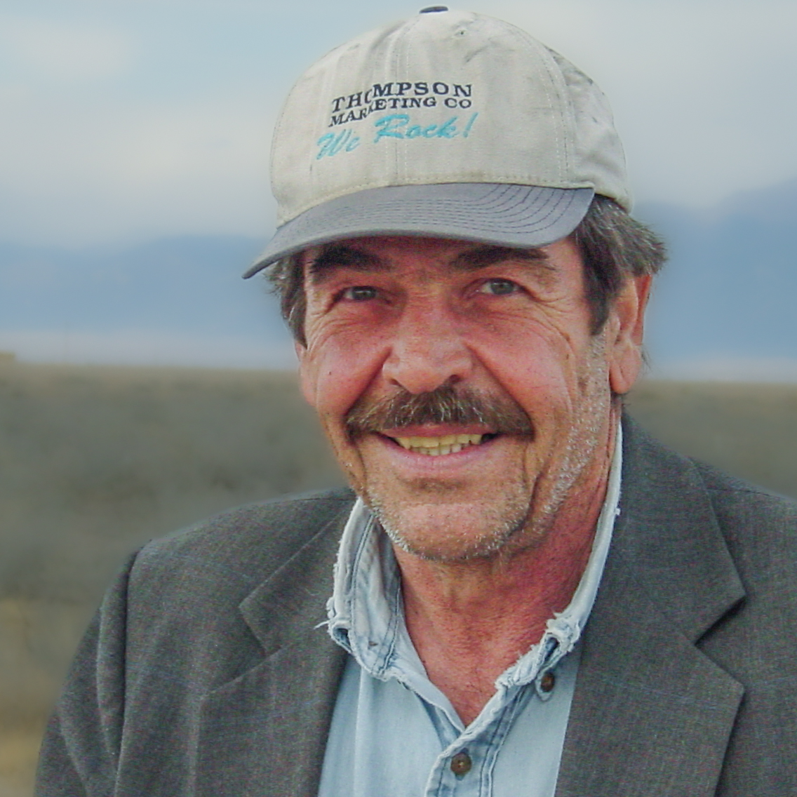 Man smiles wearing a sport coat and ball cap. 