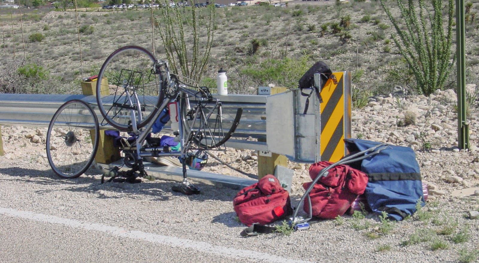 Bags strewn on the side of the road with a bicycle upside down leaning on a guardrail. Desert surrounds. 