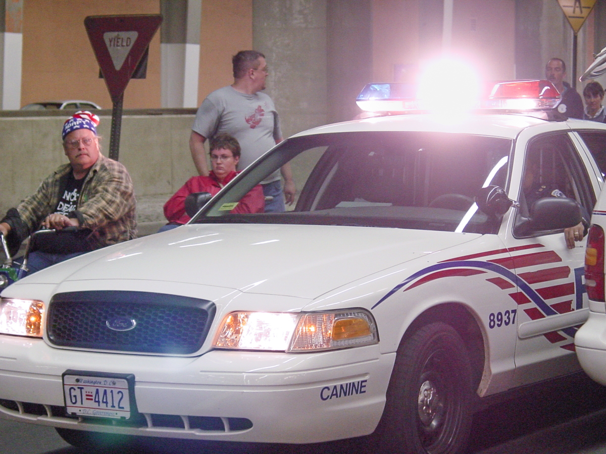 A police car with flashing light is in front of a man using a wheelchair 