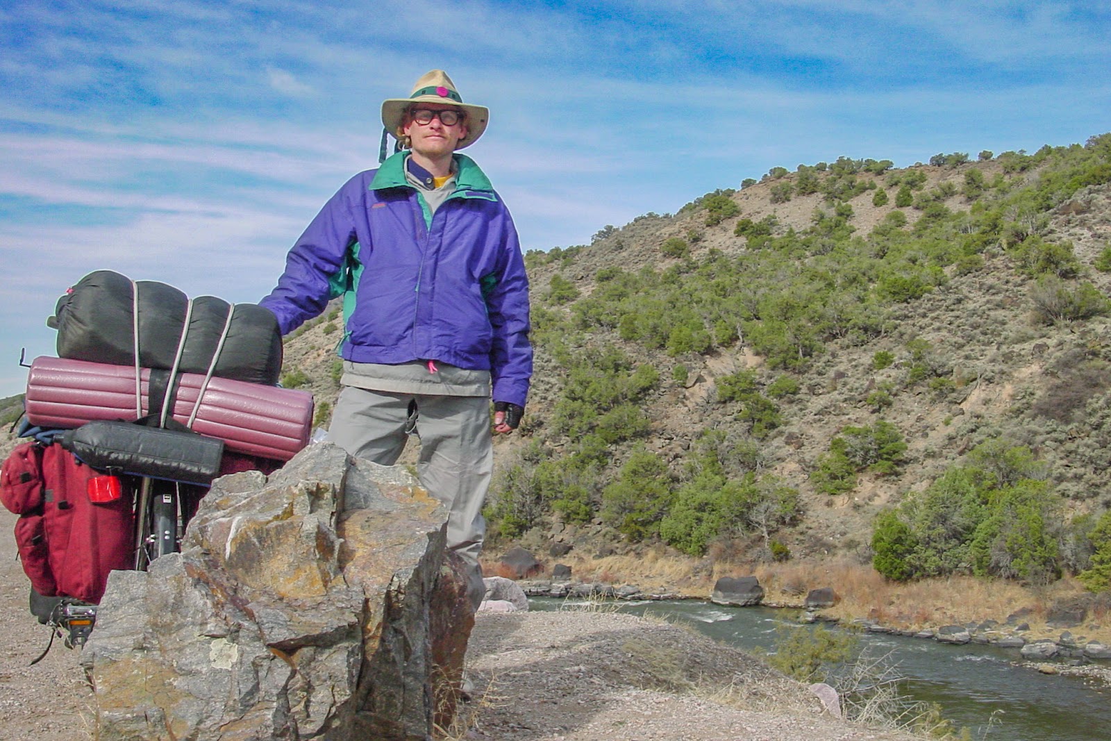 Man in wide-brimmed hat stands by a bicycle loaded with camping gear. A river flows past and behind.