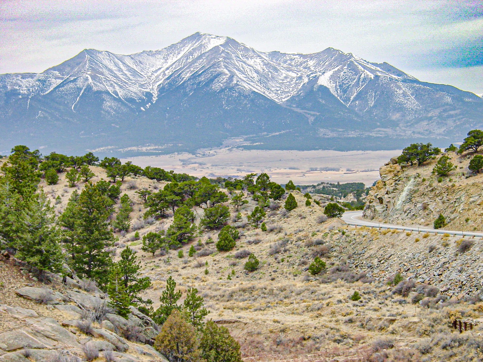 A very tall mountain with snowy peak, among other mountains. A road winds down to the valley in front of the mountains. 