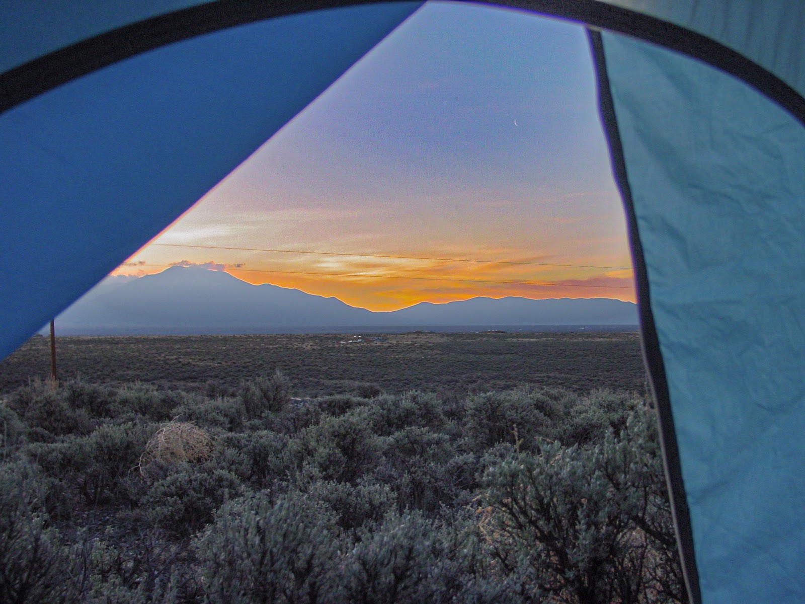A tent opening frames distant mountains silhouetted against a yellow sky. Dark desert shrubs are in the foreground. 