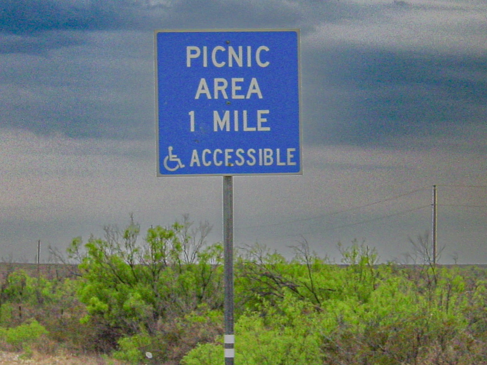 A sign against a cloudy sky that says: PICNIC AREA 1 MILE ACCESSIBLE and includes the universal access symbol. 