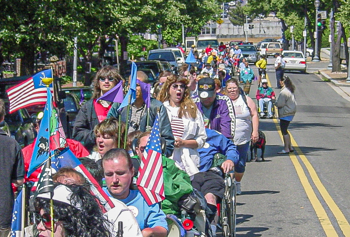 Colorful line of people with flags and open mouths take up a full lane of traffic. 