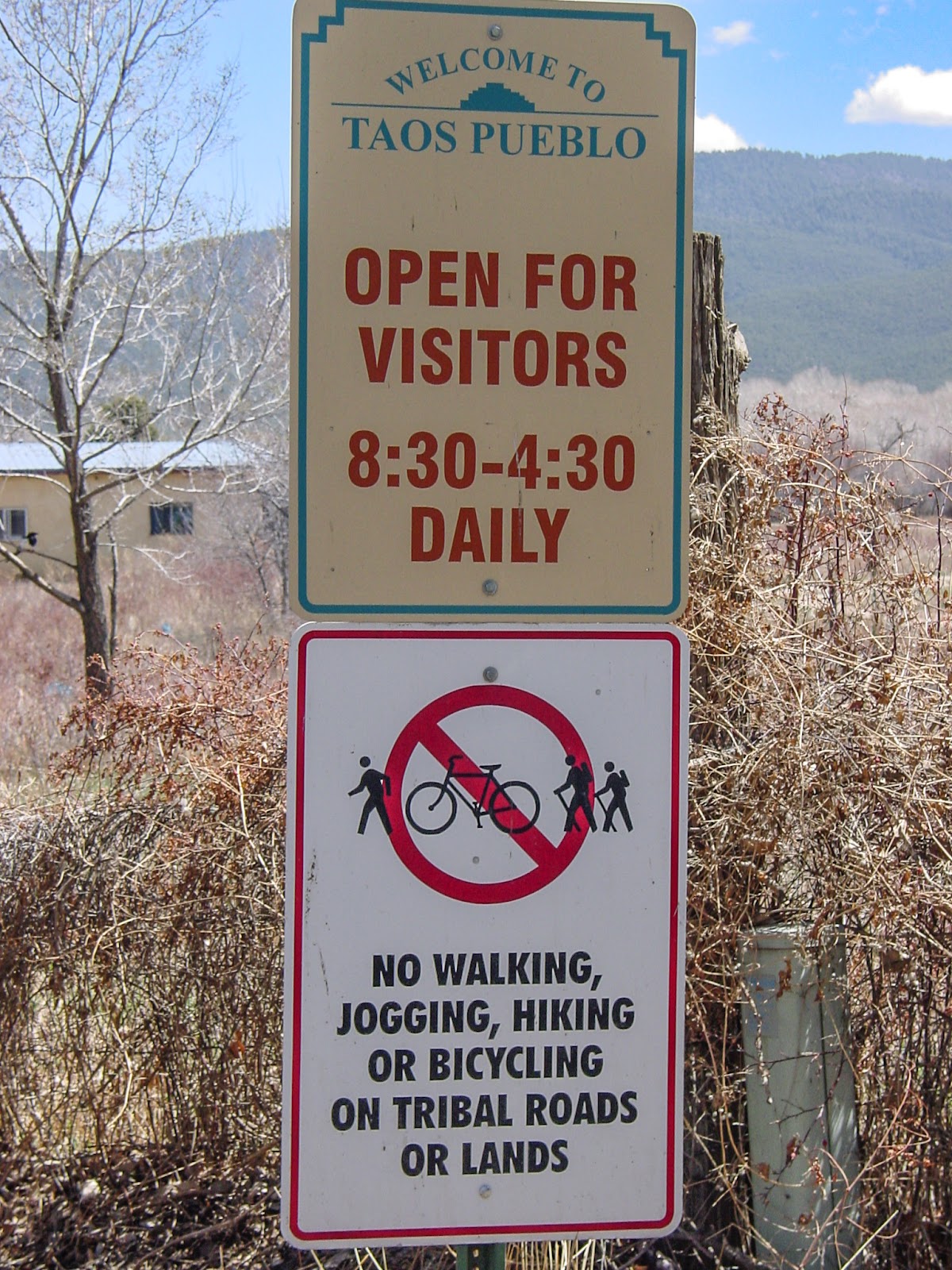 Two signs, the one on top says: WELCOME TO TAOS PUEBLO. OPEN FOR VISITORS 8:30 - 4:30 DAILY
The lower sign includes a no symbol through a bicycle and says: NO WALKING, JOGGING, HIKING OR BICYCLING ON TRIBAL ROADS OR LANDS. 