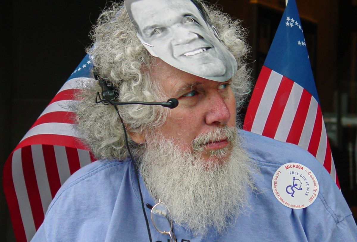 Man with white hair and beard wears a headset with microphone at his eye and a mask pushed up on his forehead. 