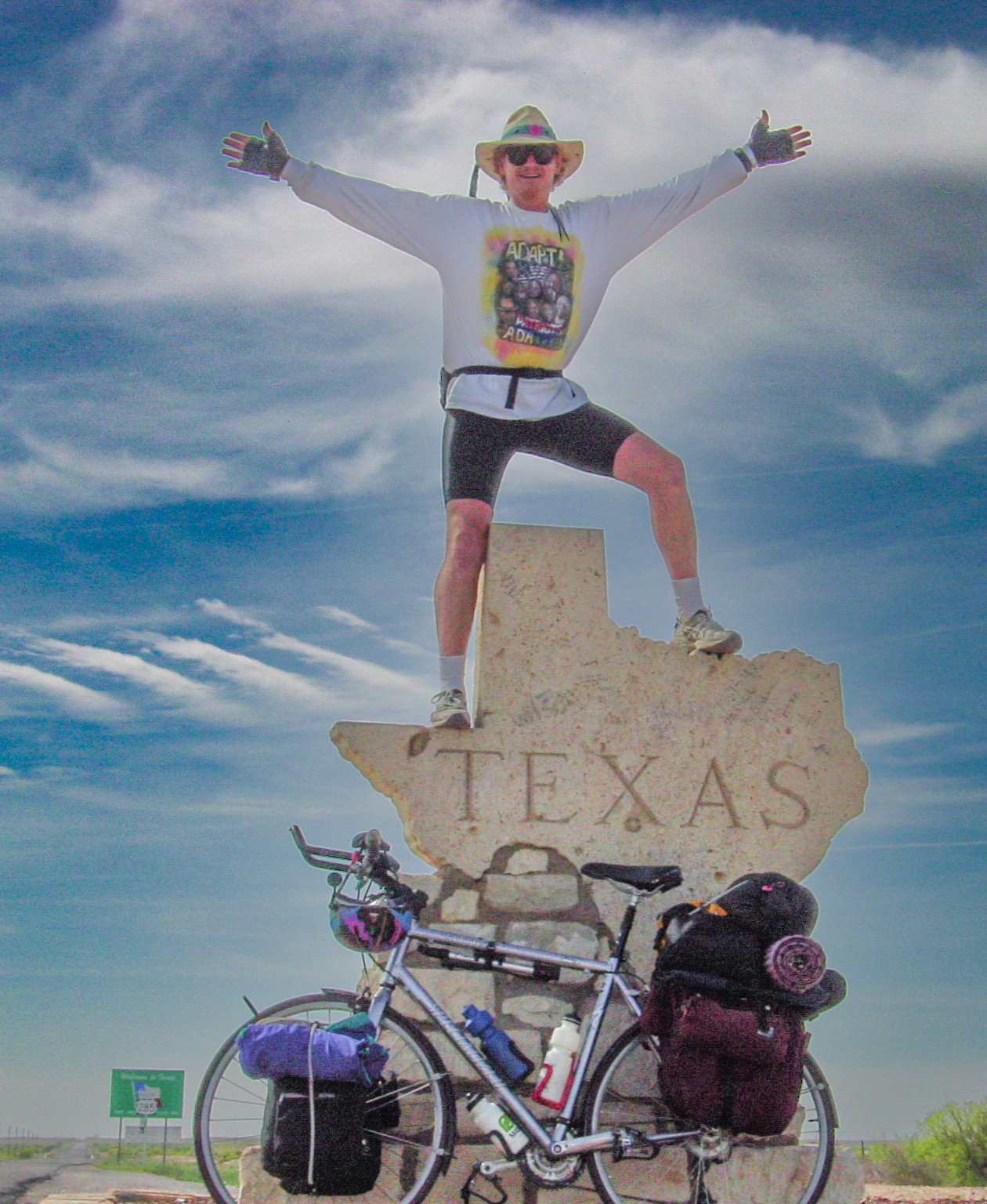 Man stands atop the Texas state line marker in the shape of the state, bicycle leans on the sign below. Blue sky with clouds in open country. The entire bike is not visible. 