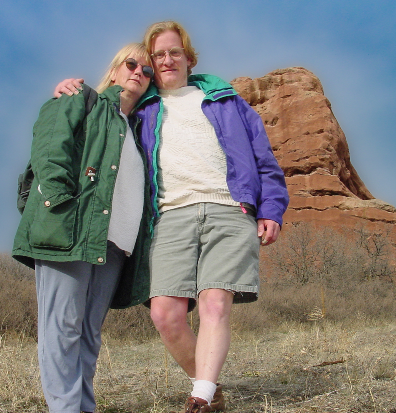 Woman in sunglasses and man in shorts with his arm around her. A reddish pointy hill is in the background. 
