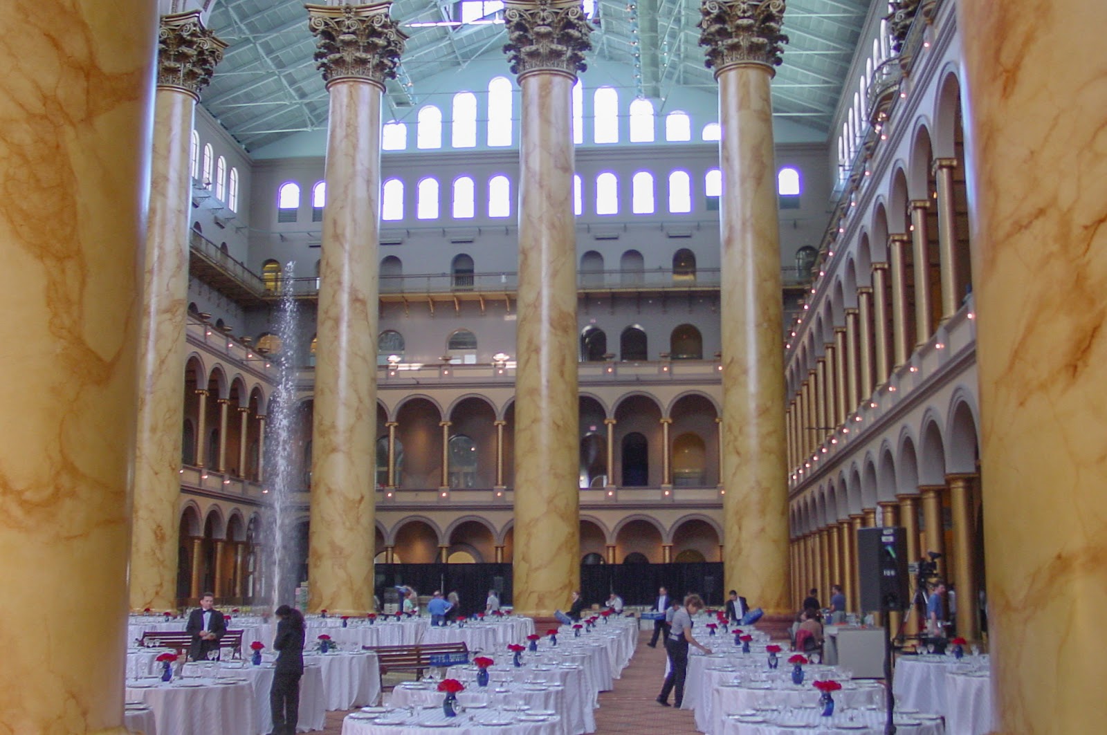 Huge open area with gigantic marble columns and people setting up tables with white tablecloths. 