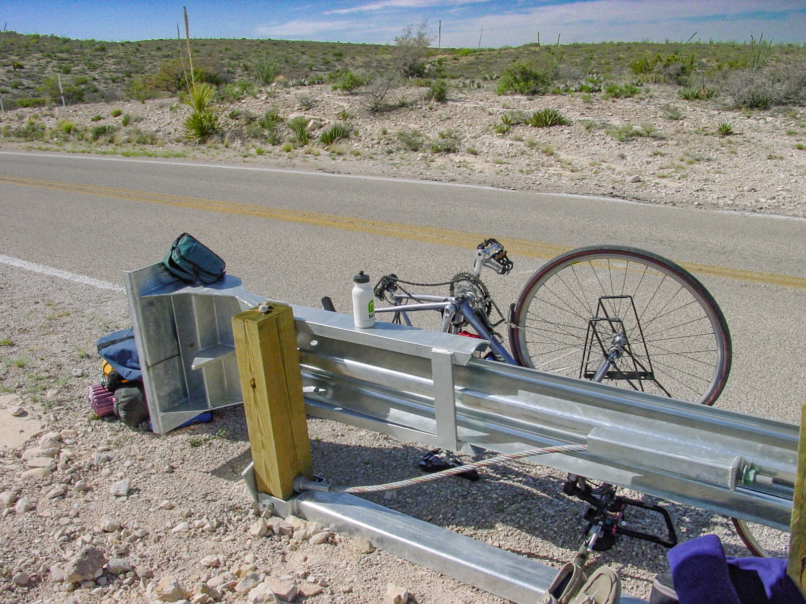 Bags strewn on the side of the road with a bicycle upside down leaning on a guardrail. Desert surrounds. 