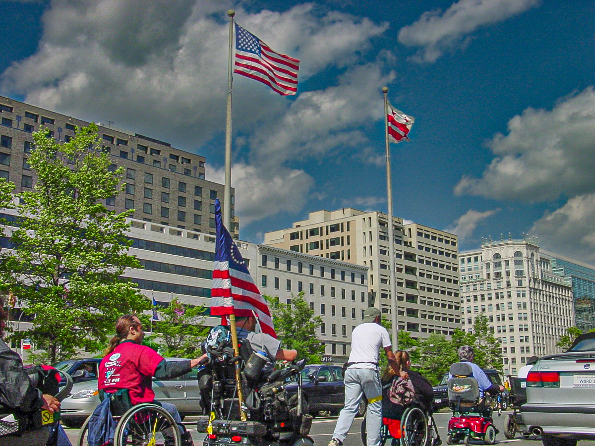 A line of people using wheelchairs march past tall buildings and blue cloudy skies. 