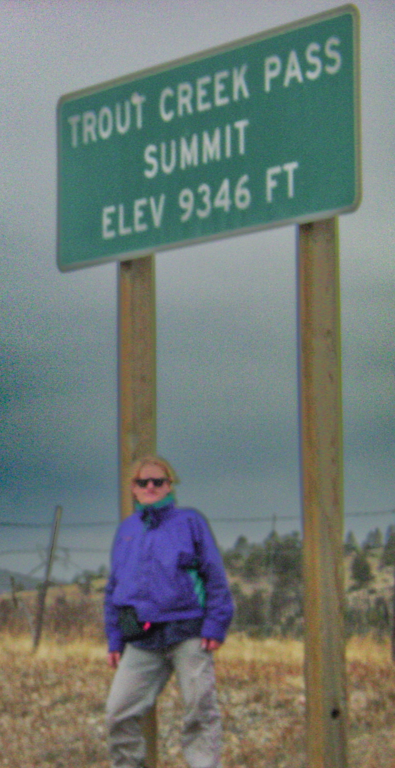 Man in coat leaning against a sign: TROUT CREEK PASS SUMMIT ELEV 9,346 FT