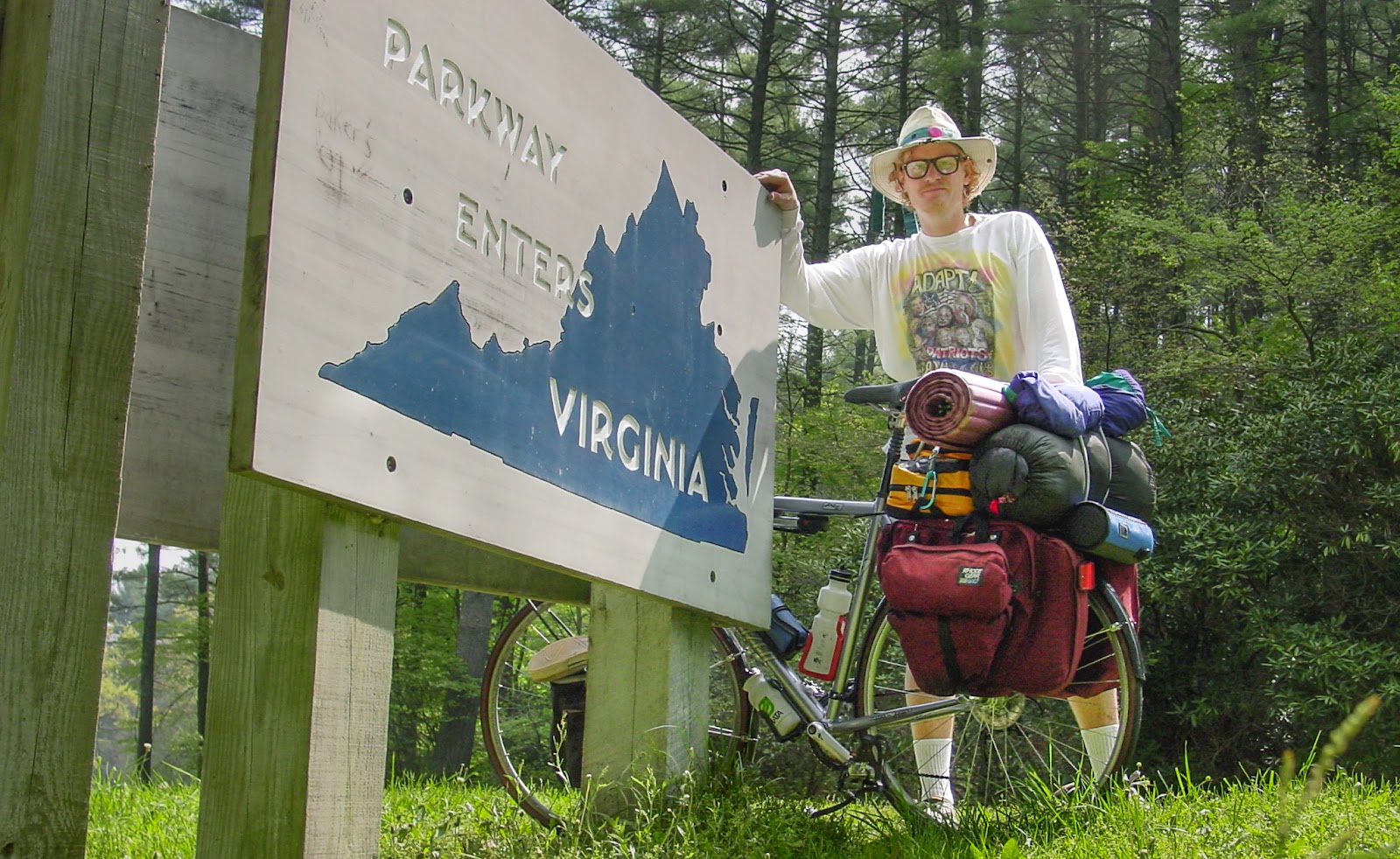 Cyclist in a wide-brimmed hat stands behind a bike and leans on the the Virginia state line marker.