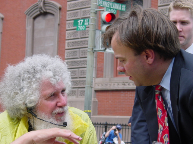 Man with white hair and beard in a yellow rain poncho speaks with a young man in a blue suit.