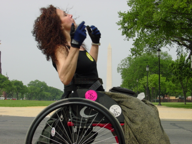 Woman using a manual wheelchair tosses her head back wearing dark fingerless gloves.
