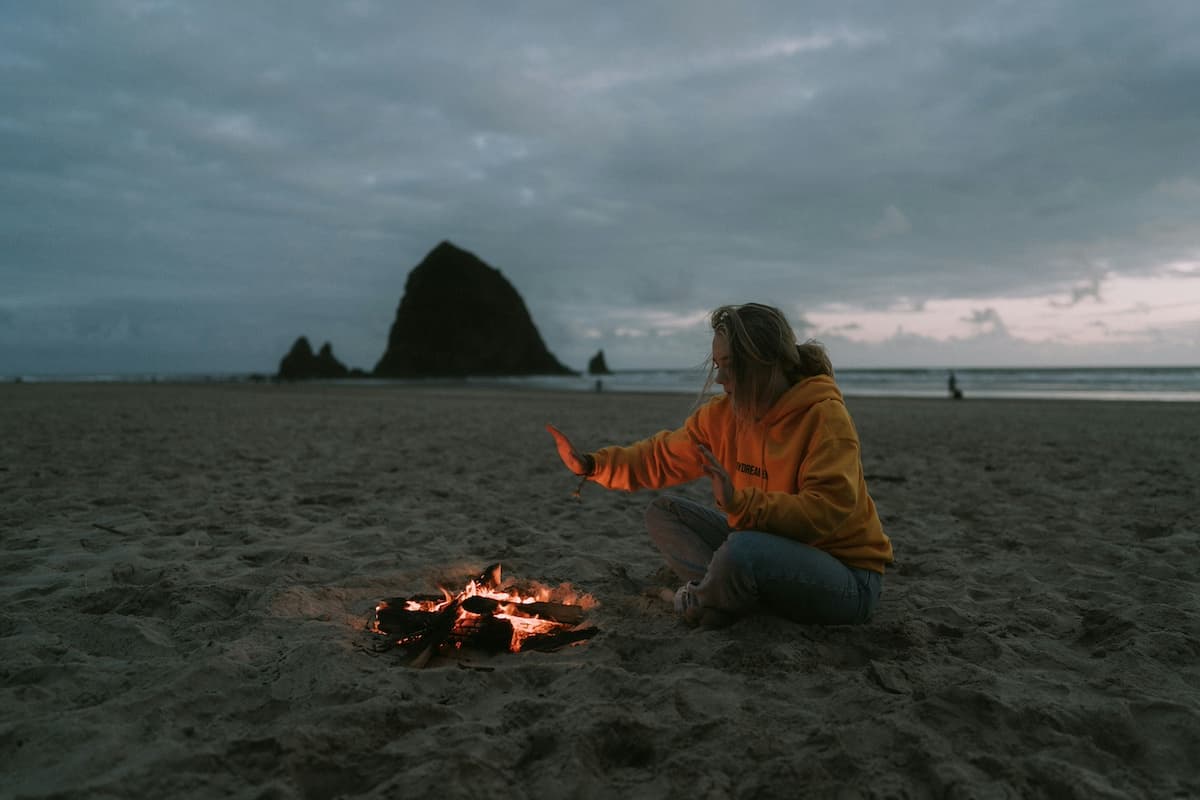 A woman warming her hands beside a fire on Cannon Beach with haystack rock in the background