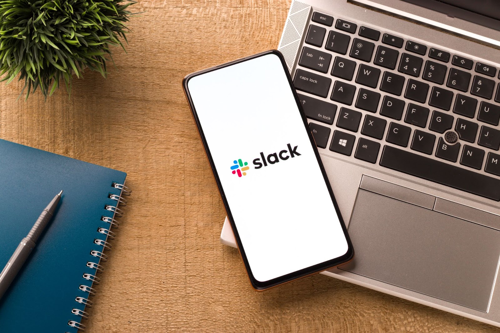 (A smartphone displaying the Slack logo rests on a laptop keyboard beside a blue notebook and pen on a wooden desk, exemplifying modern communication platforms.)