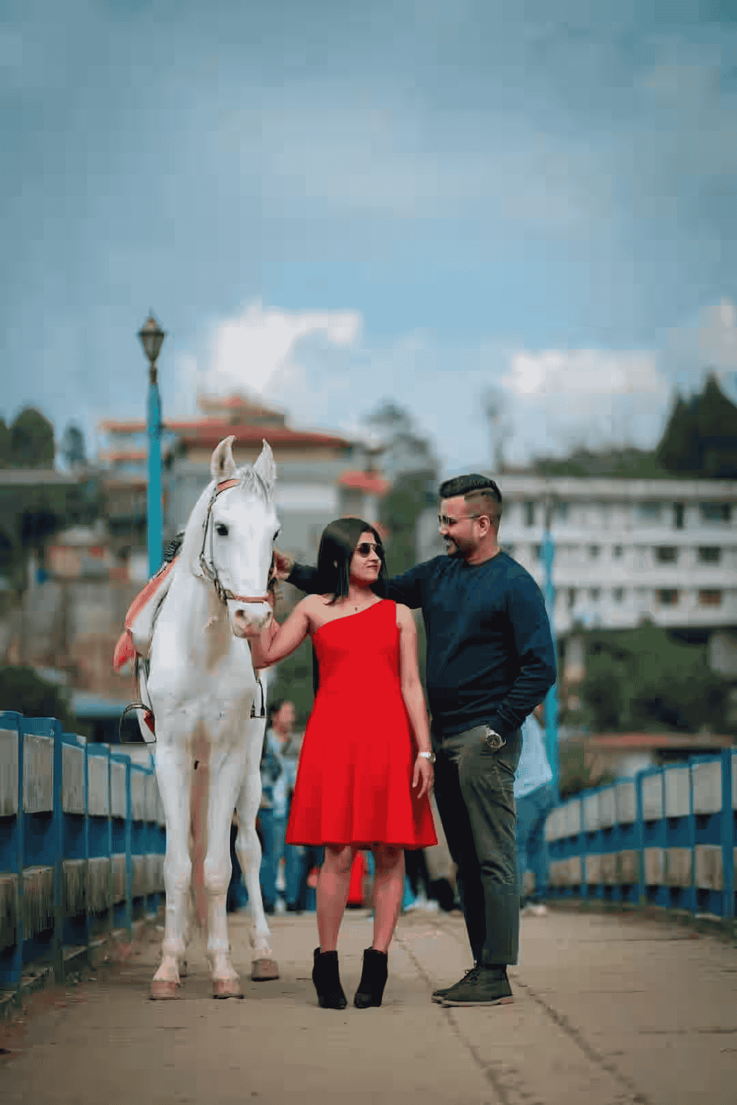 Pre wedding shoot of couple standing by a white horse, smiling and gazing into each other's eyes.