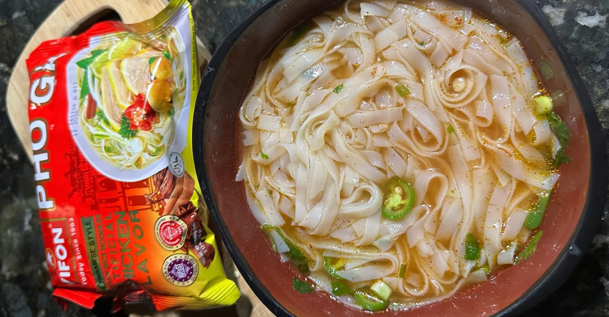 (Een kom instant kip pho met platte rijstnoedels, groene uien en gesneden chilipeper, naast een verpakking VIFON Pho Ga op een donker aanrecht.)