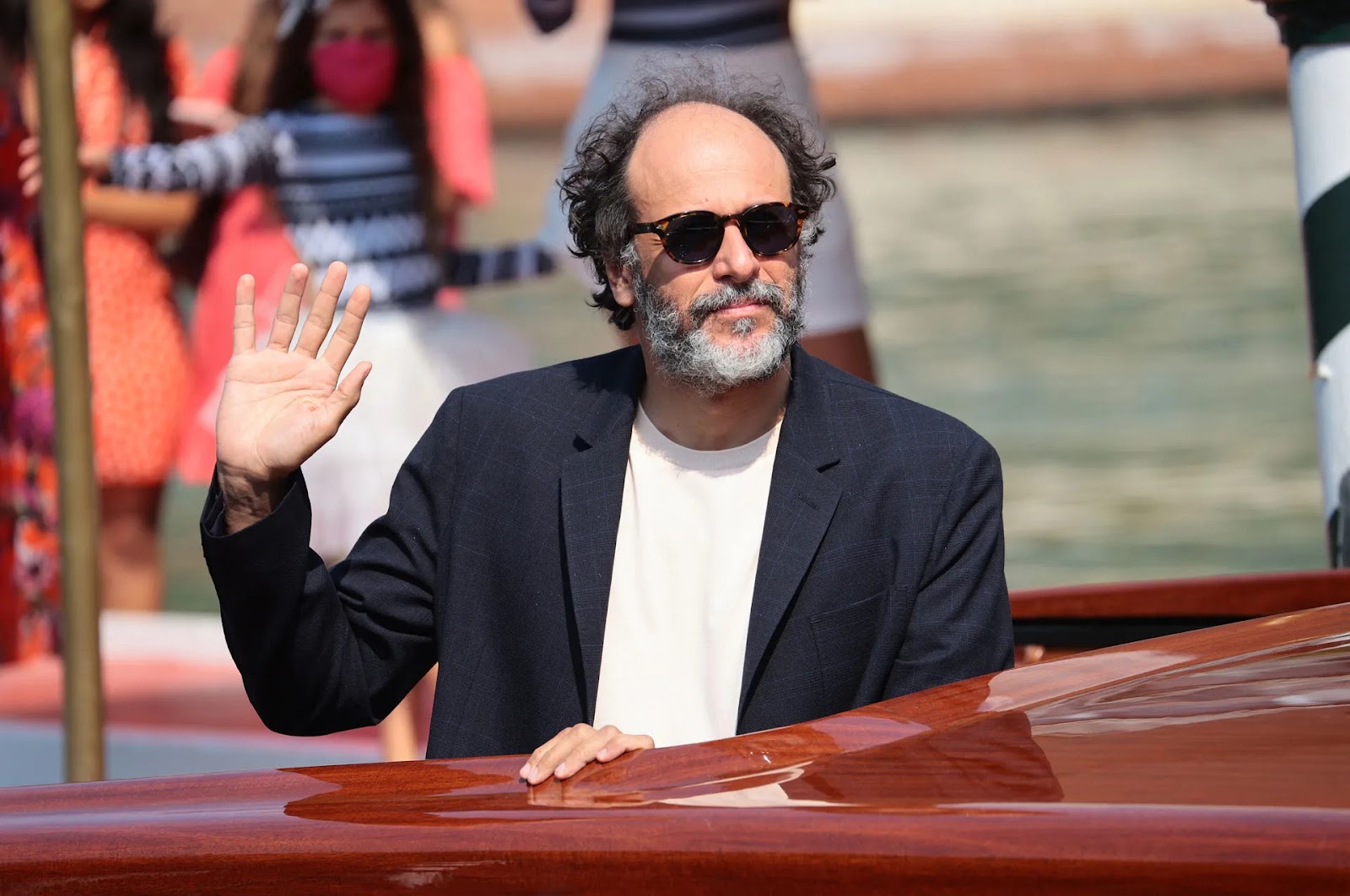 Hombre de mediana edad con barba y cabello rizado, vistiendo gafas de sol y una chaqueta oscura, saludando con la mano derecha desde un barco de madera pulida en un entorno acuático, con personas desenfocadas al fondo.