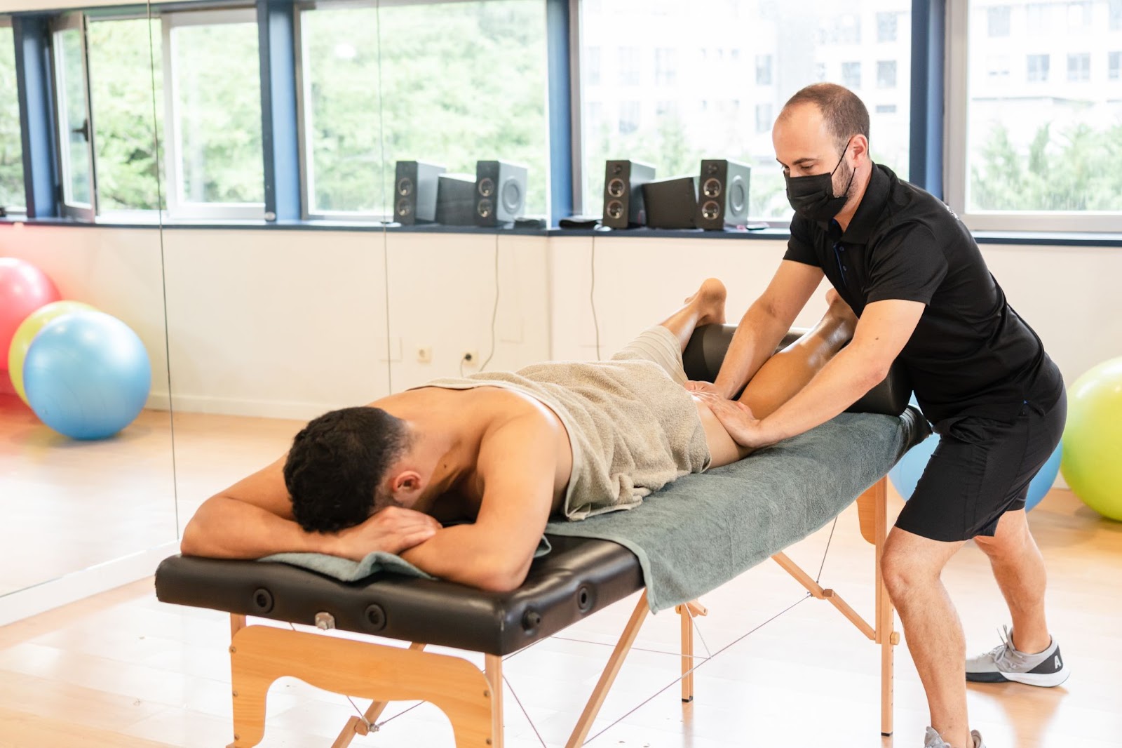 Male athlete on a table being massaged by a man in a mask with yoga balls in the background. 