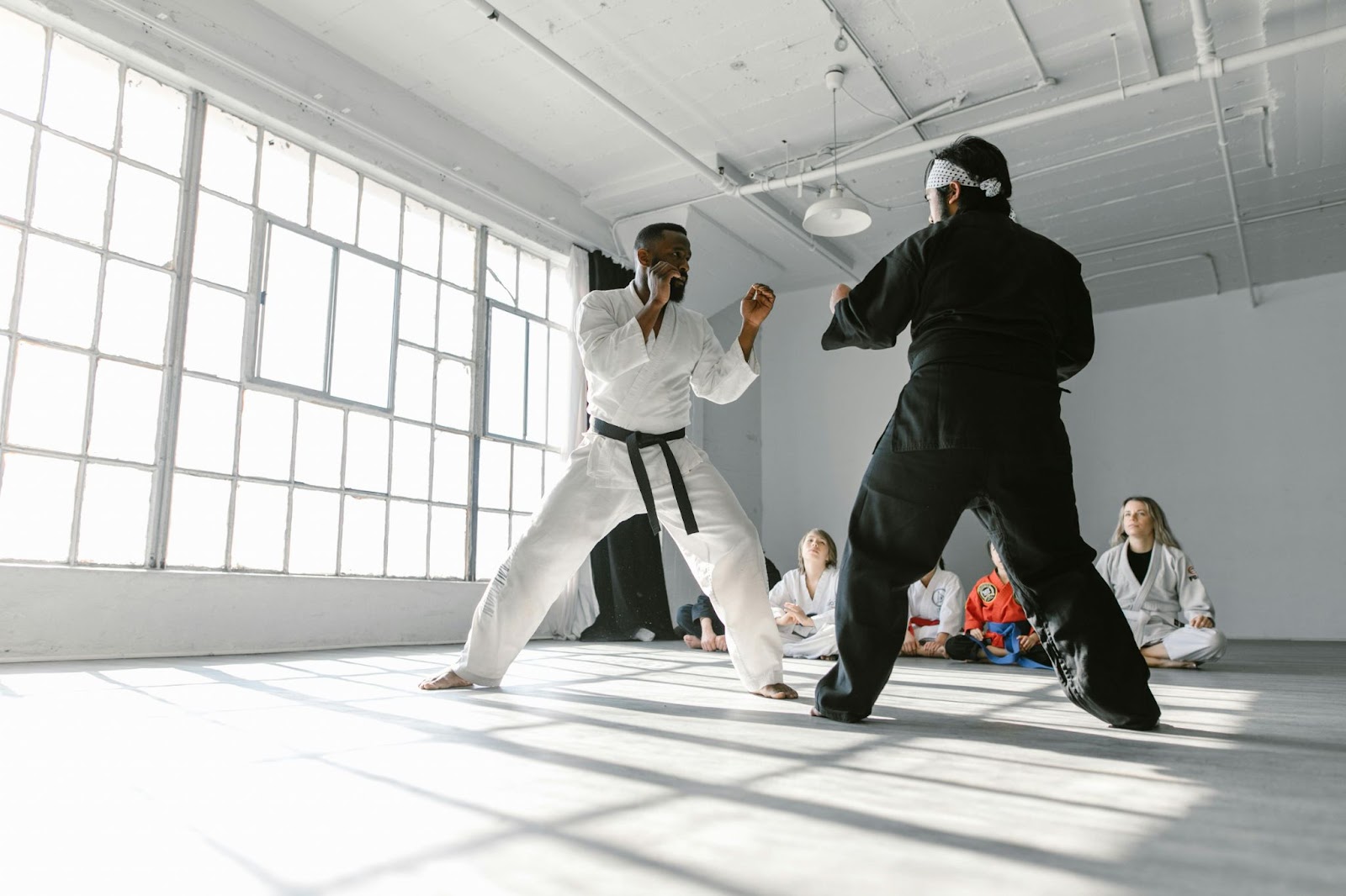 Two martial arts instructors demonstrating a stance to a group of seated students, who watch attentively from the floor in a training space.