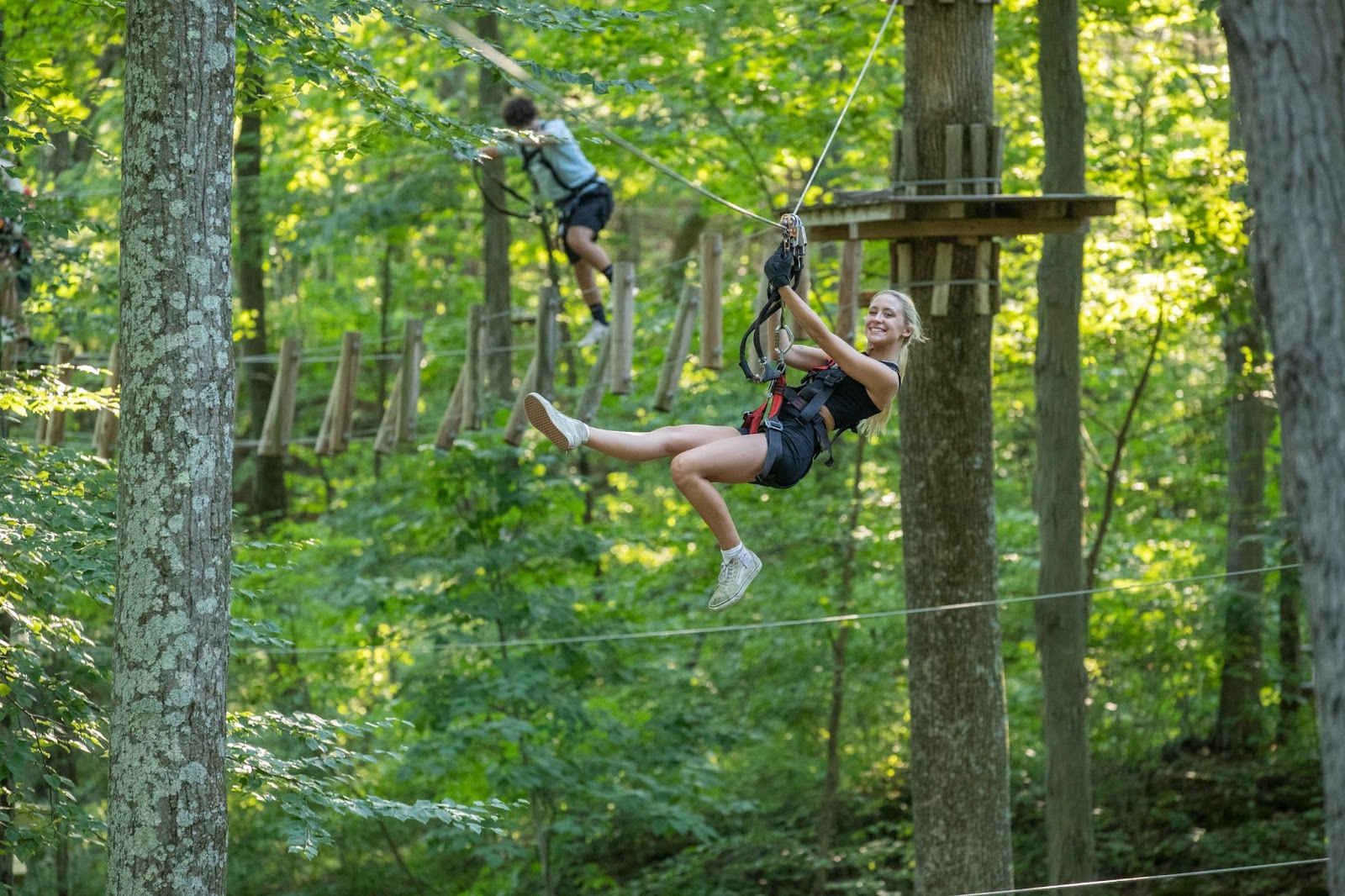 Blonde woman smiling as she navigates a zip line at TreEscape Aerial Adventure Park, Vernon, NJ, wearing safety gear and enjoying the ride as a concept of zip lining safety.