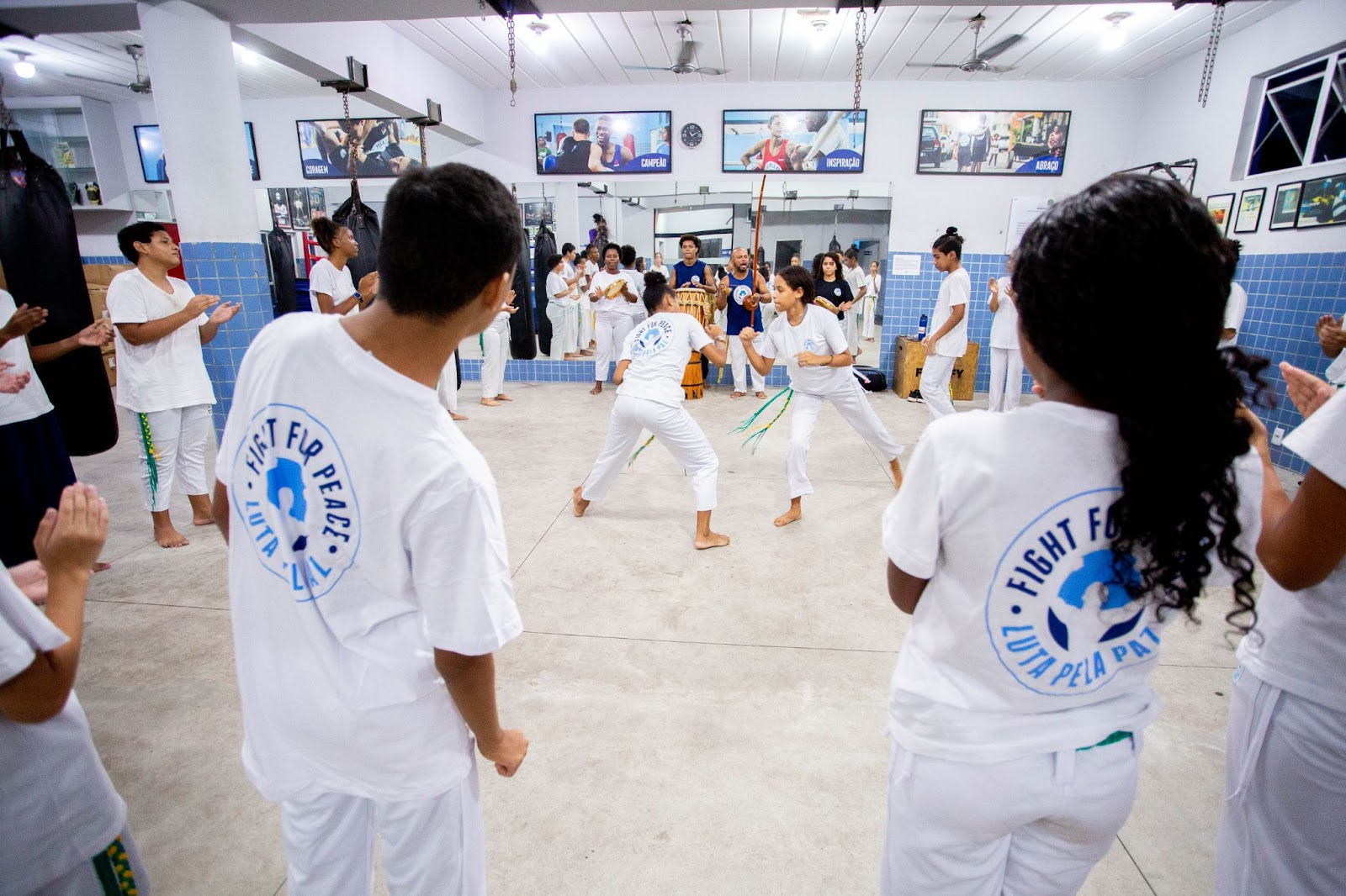 A imagem retrata uma aula de Jiu-Jitsu, onde dois praticantes estão envolvidos em um combate no tatame. Ambos usam kimono preto. Ao fundo, um instrutor veste um kimono branco e observa atentamente, enquanto outros alunos estão sentados ao redor do espaço, assistindo à demonstração. A iluminação é boa e a atmosfera é de concentração e seriedade. 