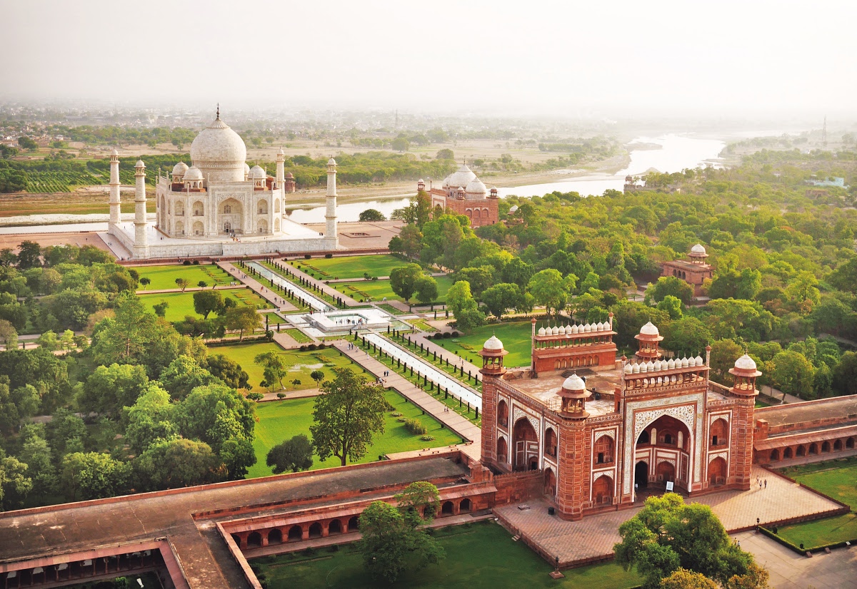 Aerial view of the Taj Mahal, an architectural masterpiece in Agra, India. The white marble mausoleum is embraced by lush gardens, a red sandstone gateway, and the Yamuna River in the background.