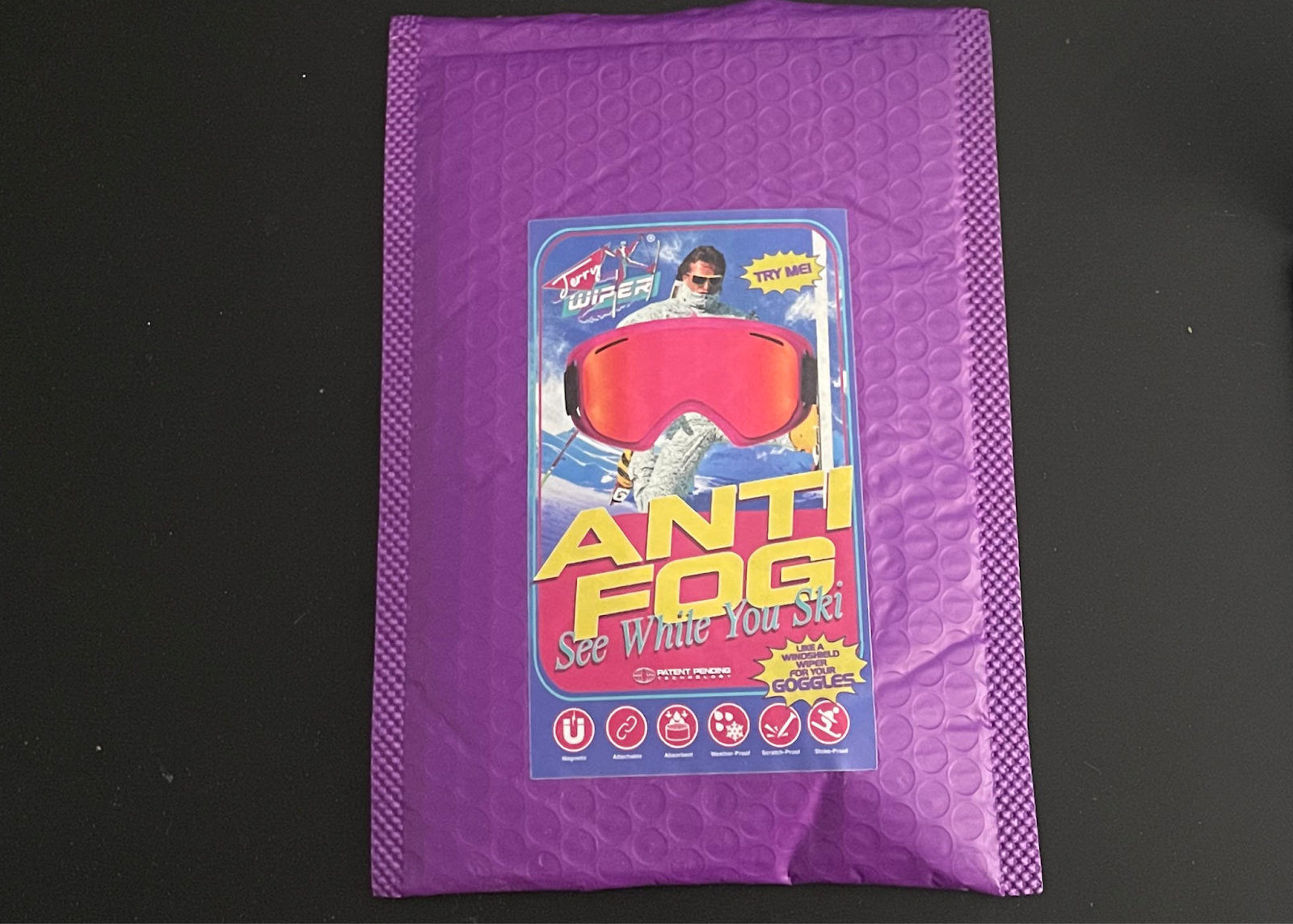 an image of the packaging for the envelope the Jerry Wiper comes in. It is a purple a royal purple envelope with a picture of a retro looking skier on there. The envelope on the bottom of the page says Anti Fog in big letters, then See While you see in smaller letters. then says like a windshield for your goggles the highlights the following features: magentic attachable, absorbent, weather-proof, scratch proof, stoke-proof