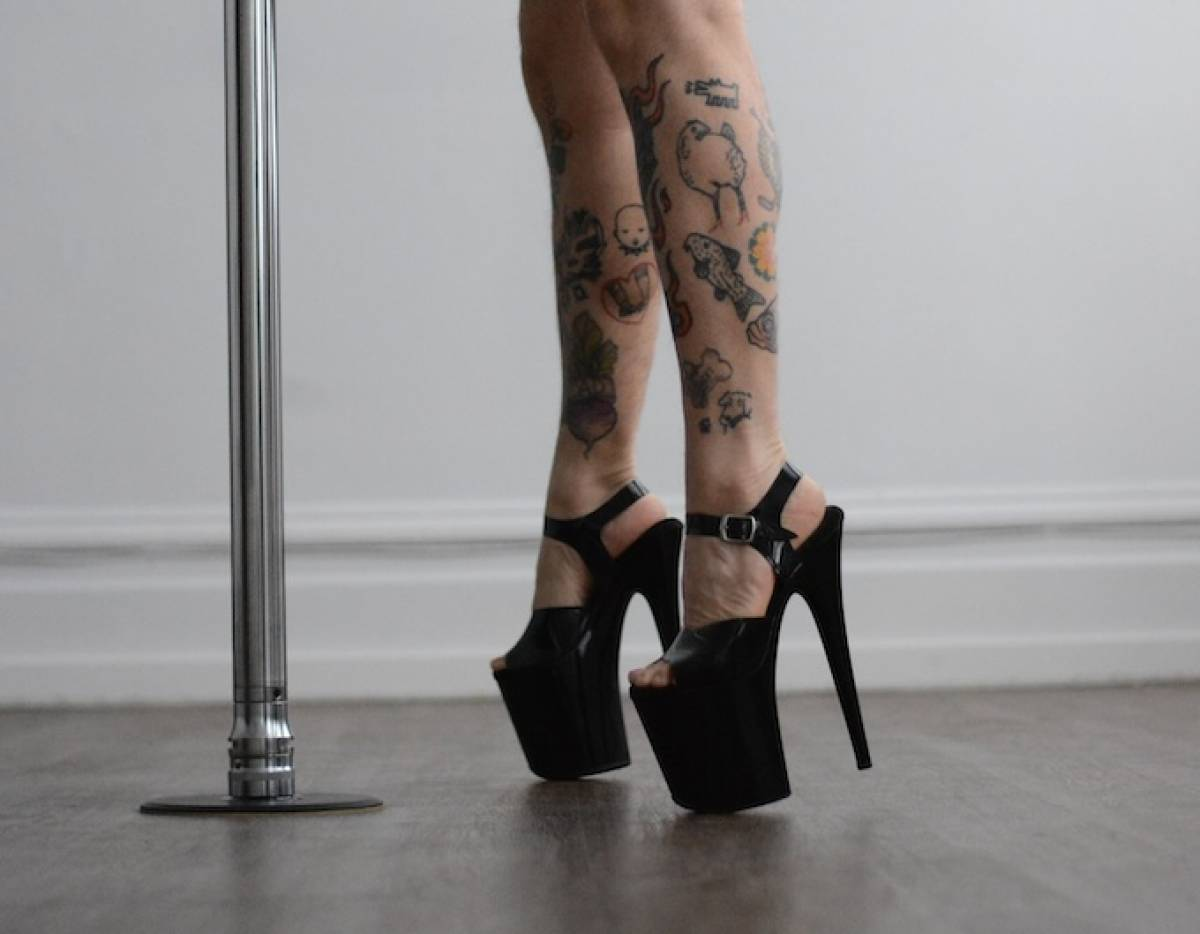 A pair of tattooed legs in black pleaser high heels, next to a pole