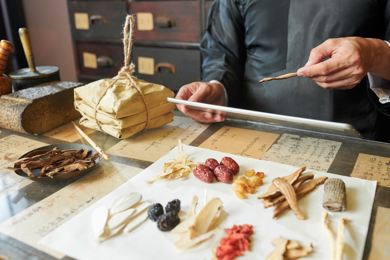 A Chinese medicine practitioner selecting herbal ingredients laid out on a clean white cloth and in a gray bowl, with a stack of herbs wrapped in paper beside her.