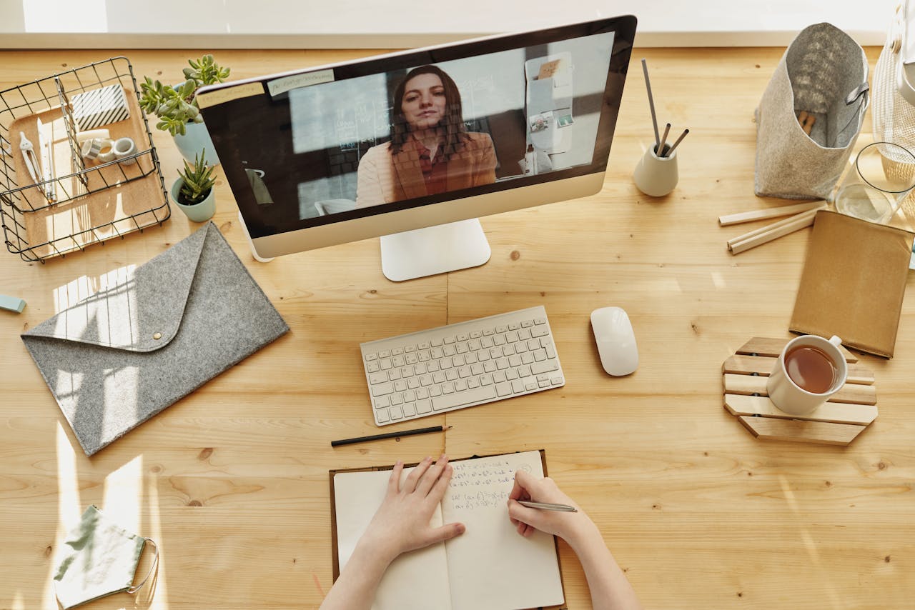 A home office setup showcasing how to create online courses, featuring a woman on a computer screen giving a lecture and another person taking notes at a wooden desk