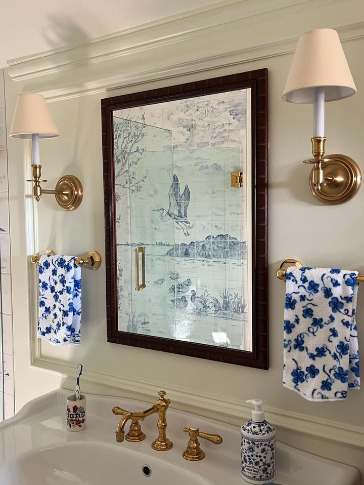 A bathroom vanity with brass fixtures, a large mirror flanked by gold sconces, and blue and white floral towels. The mirror's reflection reveals a tile mural of a heron in flight.