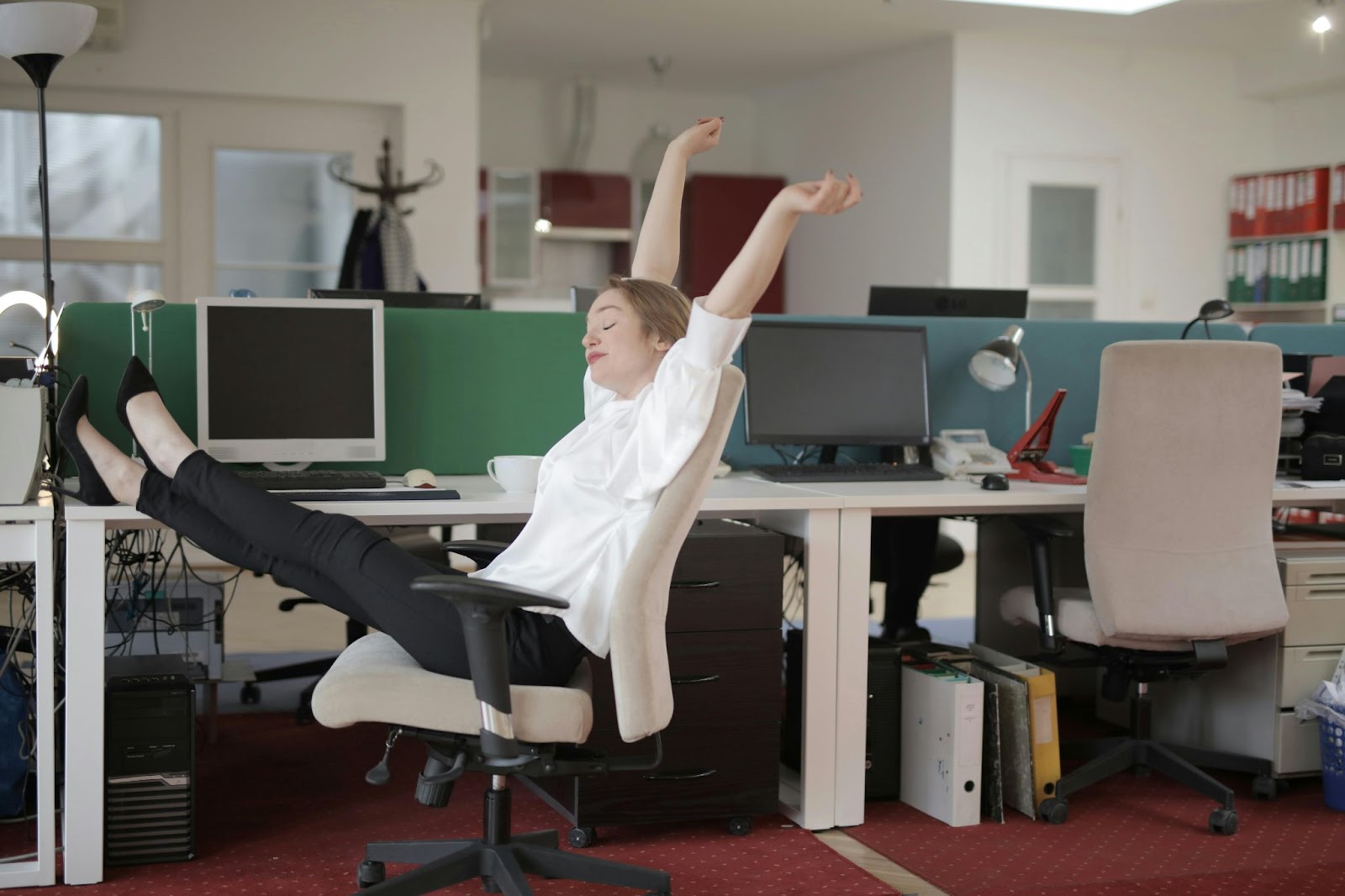A woman dressed in office attire stretches out on her chair, feet up on the desk, relaxed.