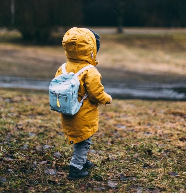 A child wearing a yellow coat and backpack walking in grass