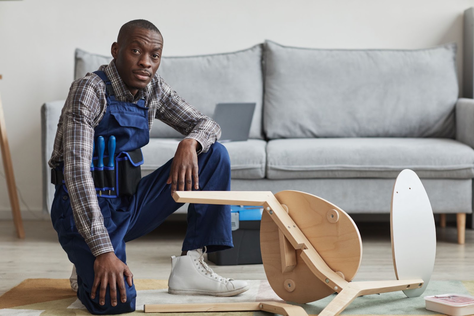 A portrait of a handyman sitting beside a broken chair for repair in front of a gray sofa.