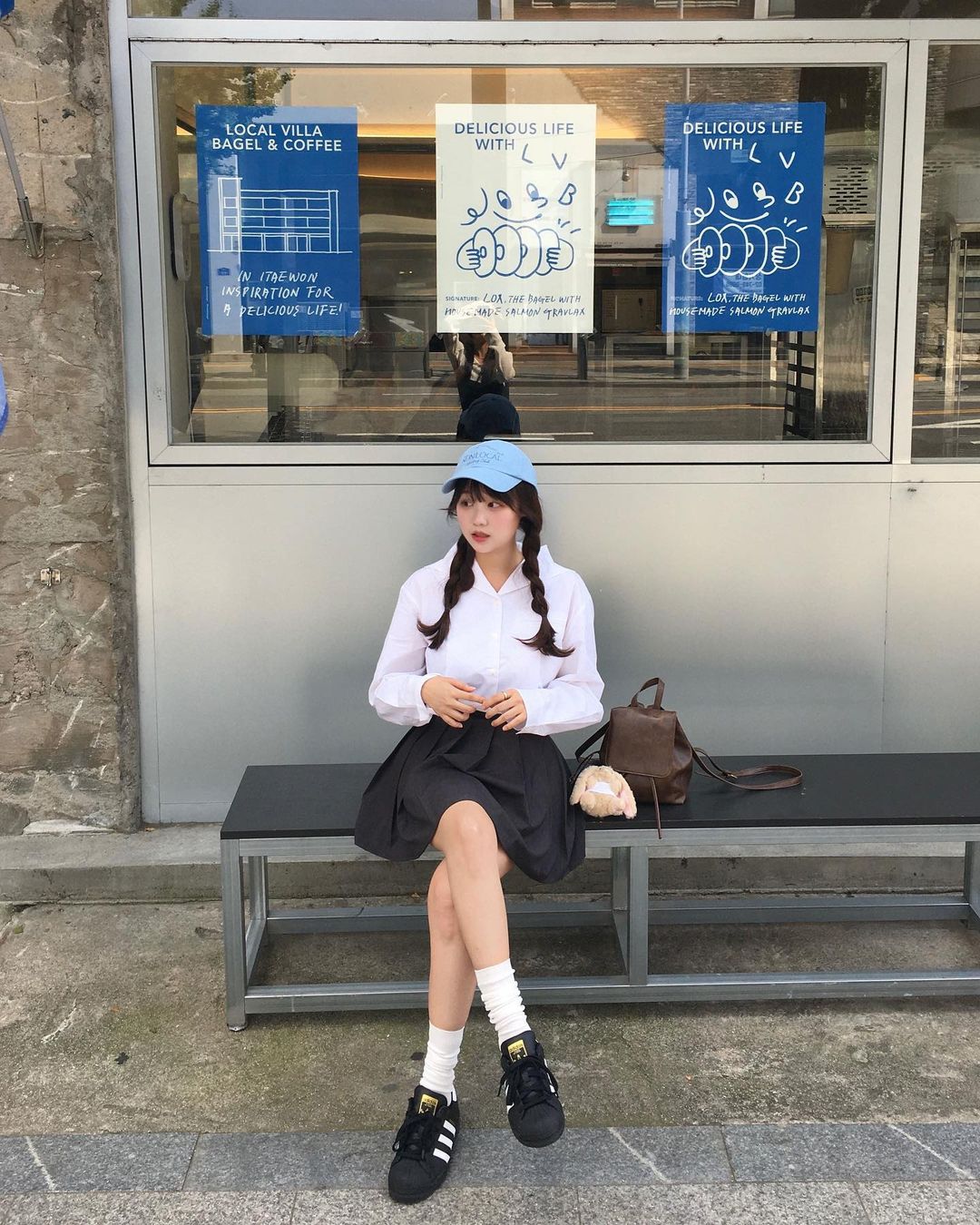 Jovem coreana usando boné azul, camisa branca, saia preta plissada e tênis preto com meias brancas