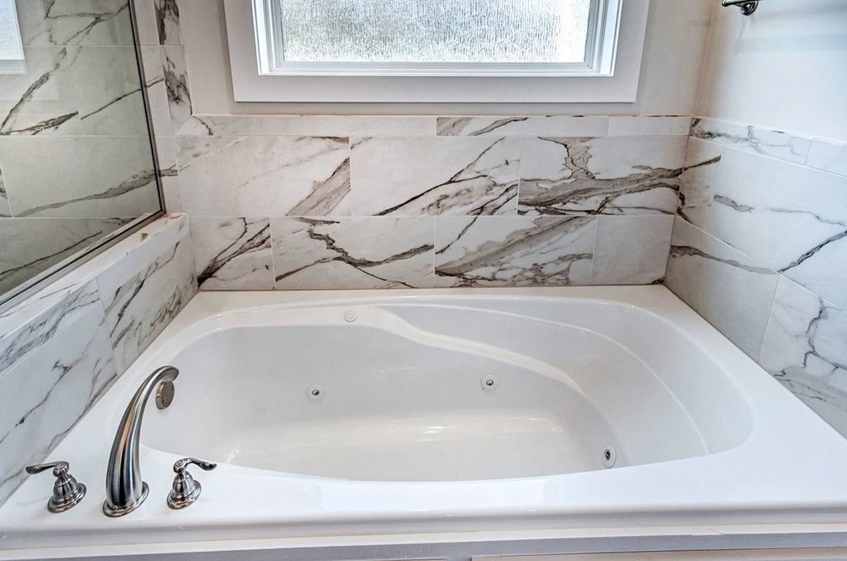 The bathtub has also chrome fixtures and a marble-tiled backsplash.