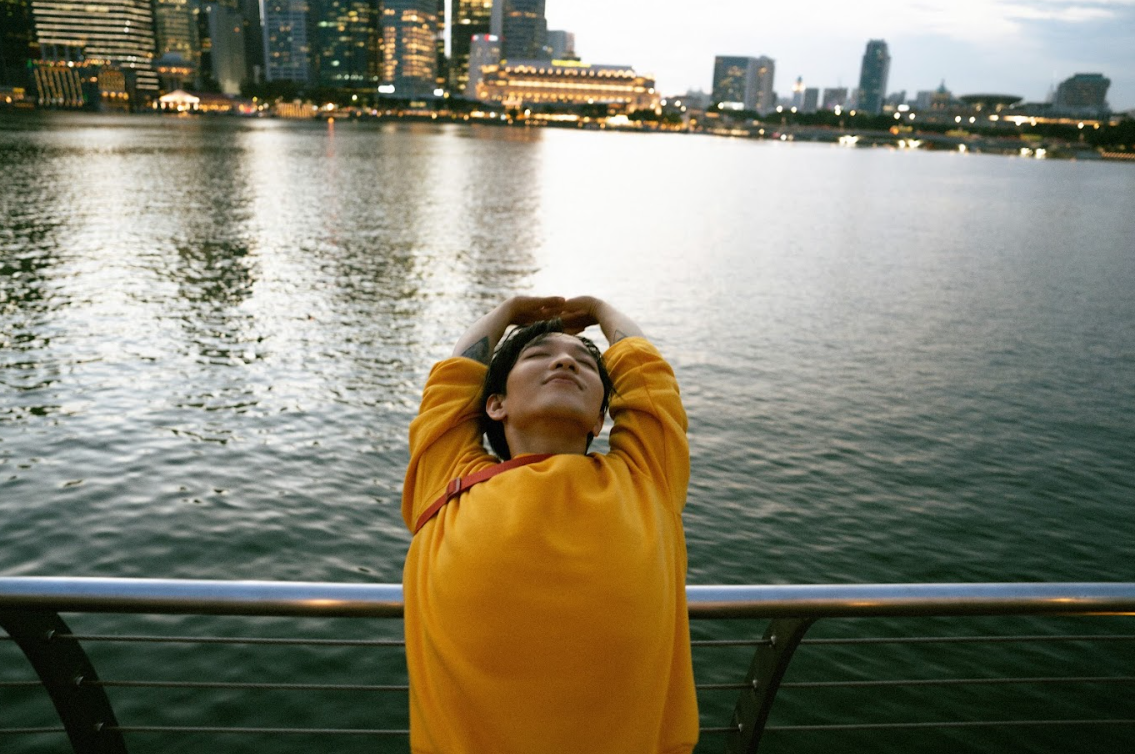 A person in a yellow shirt with his hands up in front of a body of water

Description automatically generated