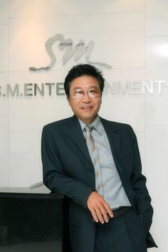 This contains a picture of  Lee Soo Man of SM Entertainment in a suit and tie standing next to a sign that says s m men's investment