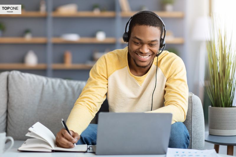 A smiling trader taking notes while learning online, preparing for a free prop firm challenge with headphones and a laptop