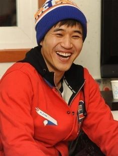 This may contain an image of Kim Jong Min sitting on top of a couch wearing a red jacket and blue hat