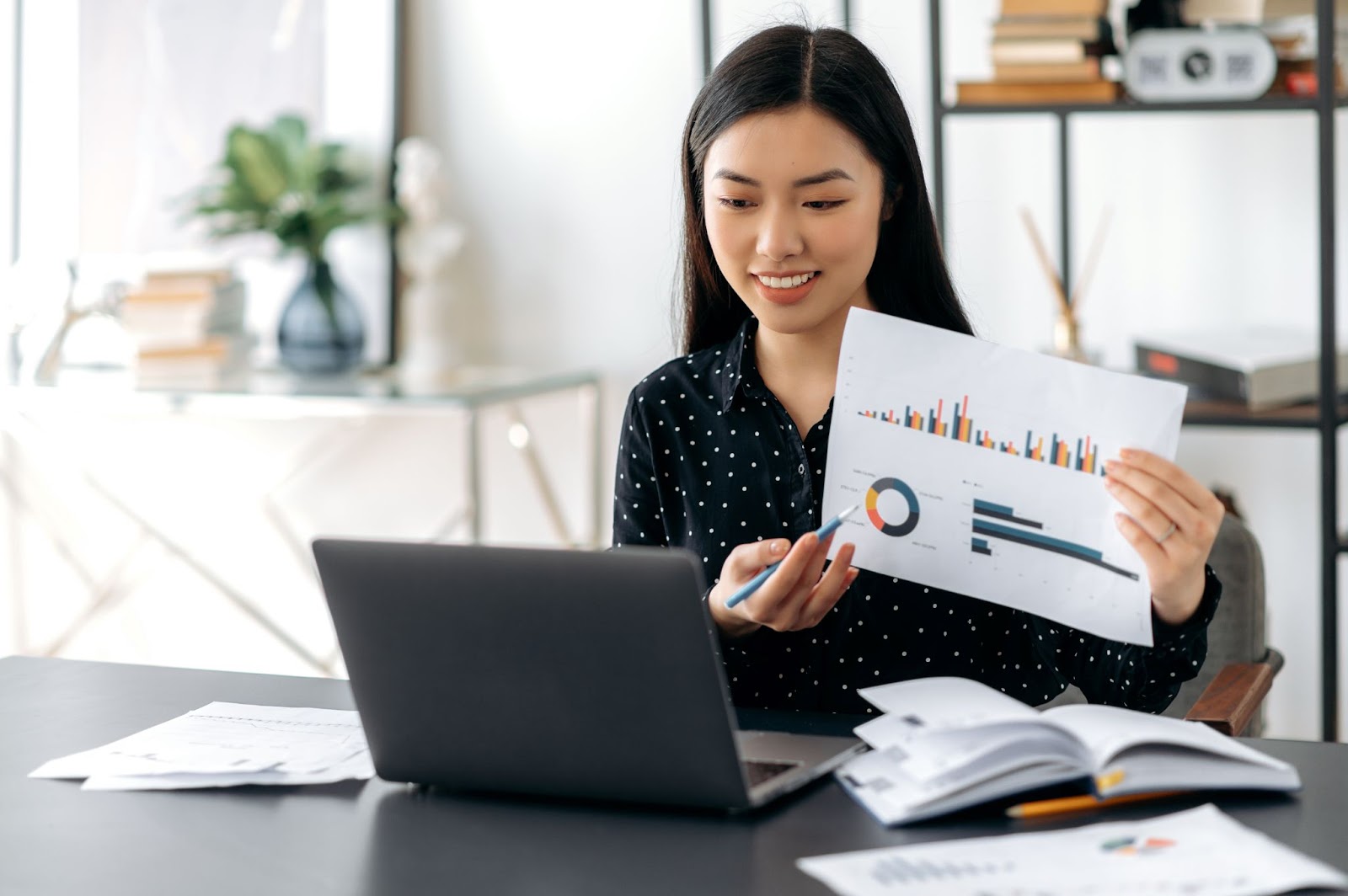 Woman holding up a piece of paper with data on it while using a laptop. 