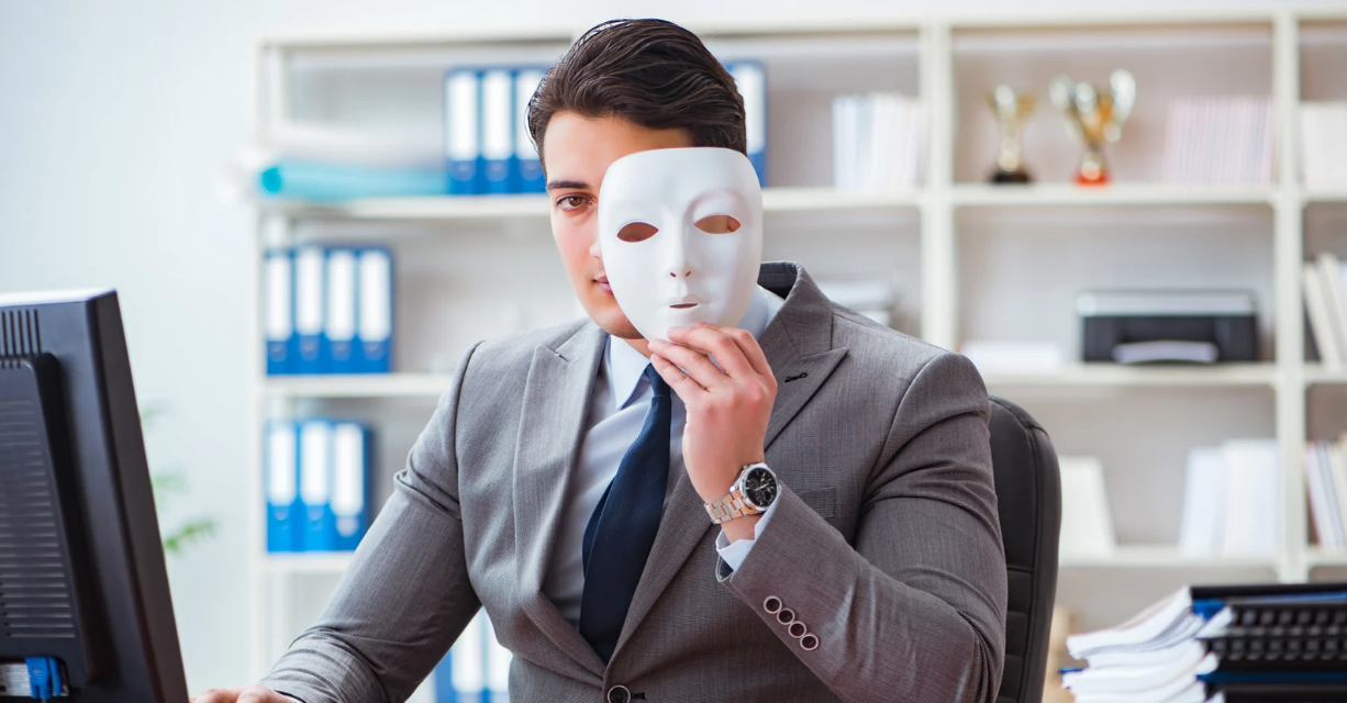 (A man in a suit sitting at a desk in an office, holding a white mask in front of his face, partially revealing his identity.)