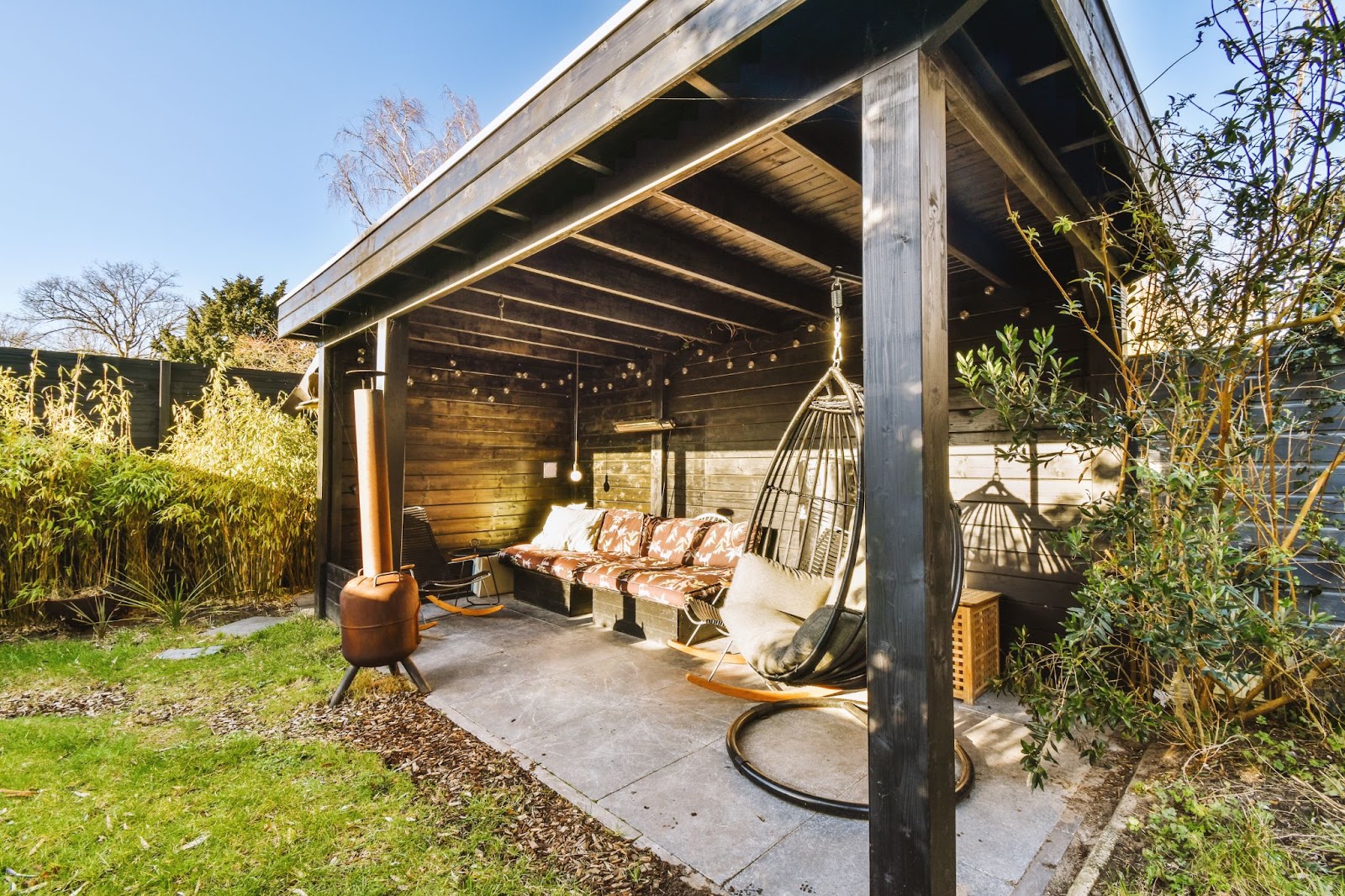 A covered patio with a solid roof, featuring a sofa, a cozy rattan egg swing, and a portable fire pit with a PVC pipe exhaust.