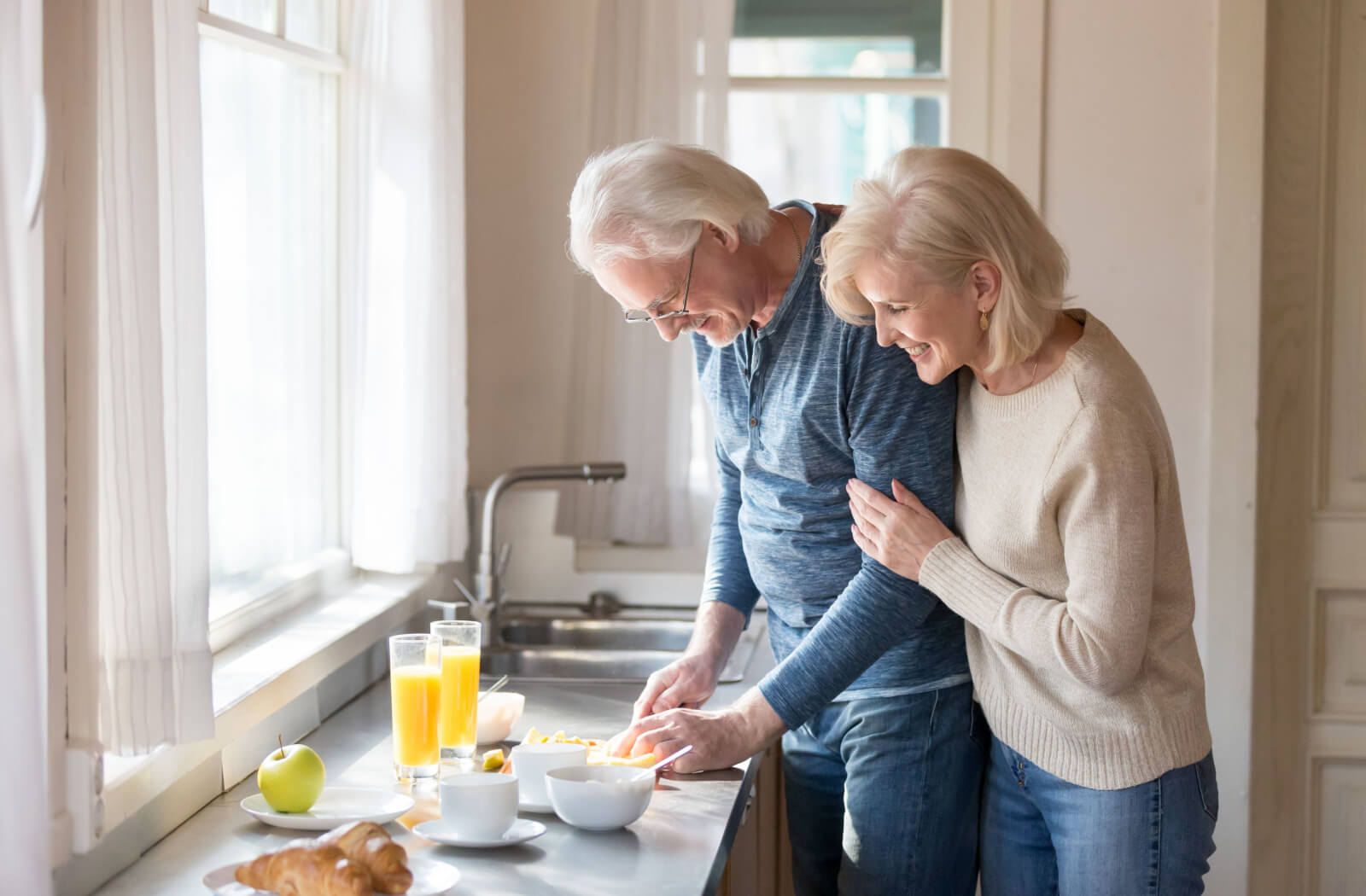 An older couple in their kitchenette cutting fruits in front of a sunlit window, making a healthy soft meal for breakfast.